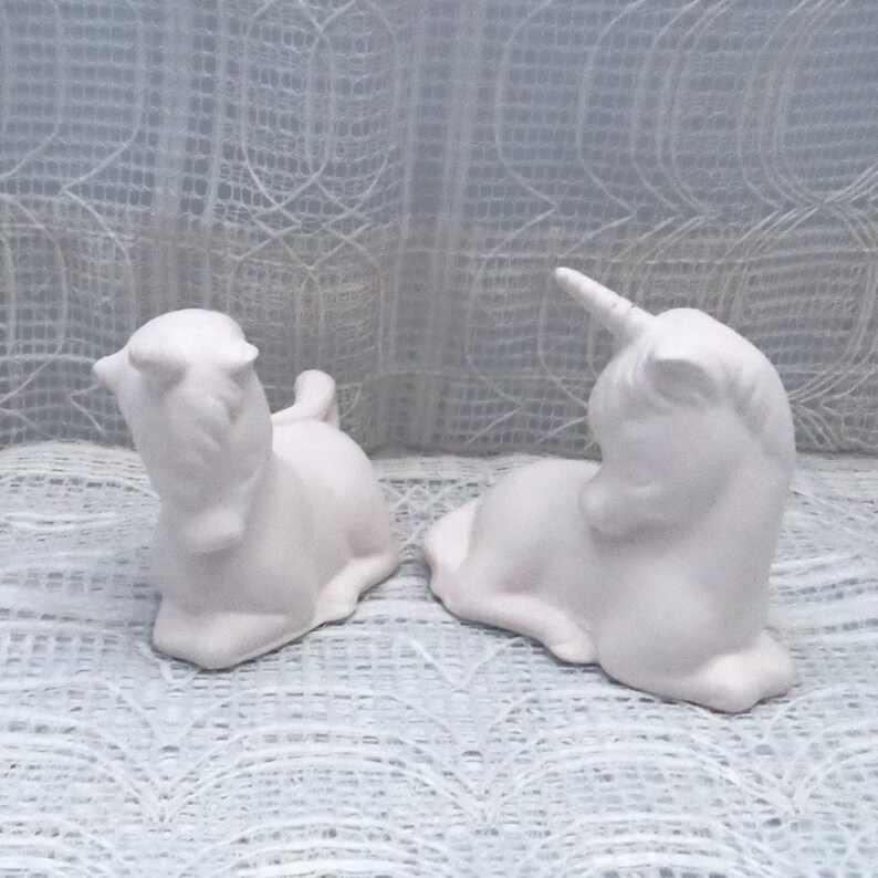 Op de afbeelding: Twee witte keramische eenhoornbeeldjes, elk met een gekrulde hoorn en een gesloten oog, staan op een wit kant oppervlak.