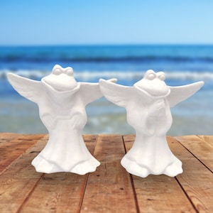 2 ready to paint ceramic frog angle standing on a wood table by the ocean.  One has his hands at his sides, one has his hands in the prayer position.