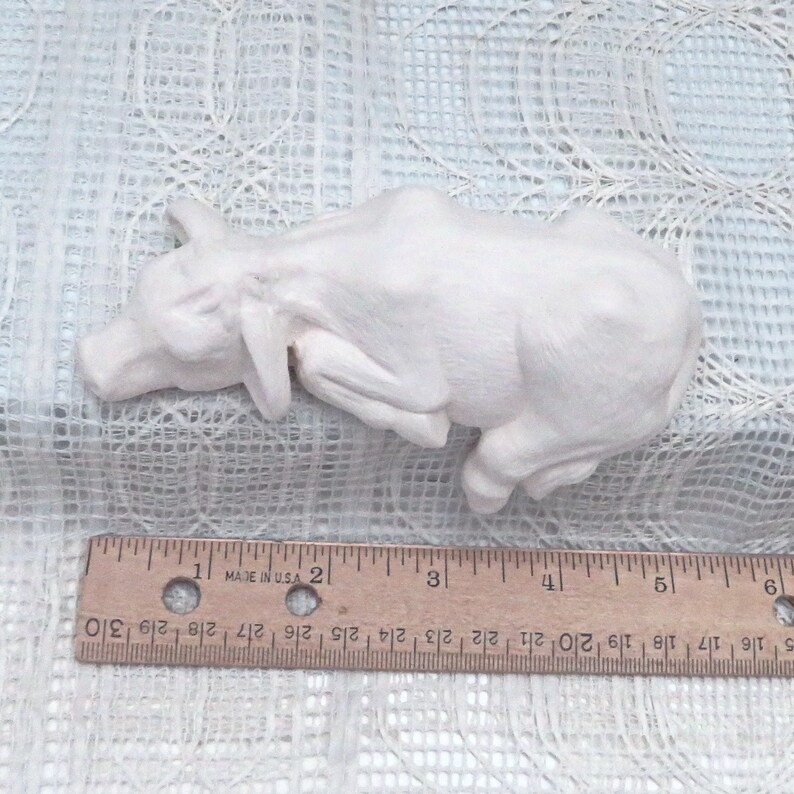May include: White ceramic figurine of a cow lying down. The cow is about 4 inches long.