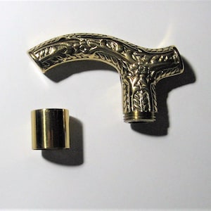 May include: A gold-colored cane handle with intricate floral and leaf patterns. The handle is curved and has a cylindrical metal piece detached. The handle is made of metal and has a shiny finish.