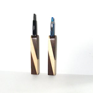 May include: Two wooden pen holders with a diagonal stripe design. One holder is brown and white, the other is black and white. Each holder has a pen inserted.