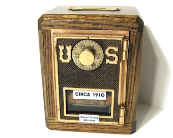 May include: A wooden bank with a combination dial and a small window. The bank has a dark brown finish with gold-colored accents. The text "UOS" is displayed on the front, along with the text "CIRCA 1910" and "Bevel Glass Window".