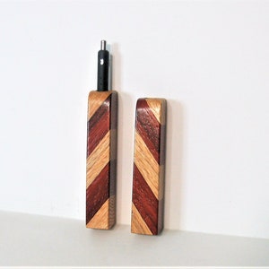 May include: Two rectangular wooden cases featuring a diagonal striped pattern of light and dark brown wood grains. One case is open, revealing a black pen with a silver tip protruding from the top, while the other case is closed and stands beside it.