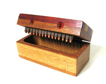 May include: A handcrafted wooden box with a hinged lid, featuring a unique design. The lid is shaped like a creature's head, with red eyes and a row of teeth. The box is made of various wood tones, creating a striking contrast.