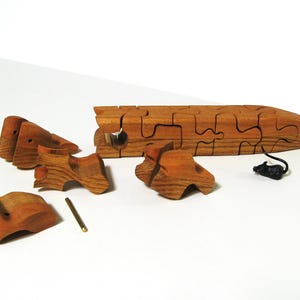 May include: A wooden puzzle in the shape of a fish, with multiple pieces that fit together. The puzzle is made of light brown wood and is disassembled, with the pieces scattered on a white surface.