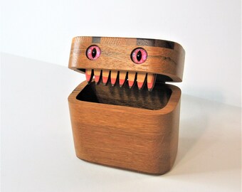 May include: A wooden box with a toothy mouth and pink eyes. The box is open and the inside is empty.