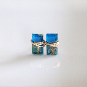 May include: A pair of rectangular stud earrings. Each earring features a vibrant blue resin block with gold wire wrapping. The earrings are set against a white background, with a gold accent visible on the left.