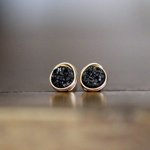 May include: A pair of gold-toned stud earrings with a dark, textured, druzy stone center. The earrings are round with a gold border, and the dark stone has a sparkling, crystalline appearance. The earrings are displayed on a reflective surface.