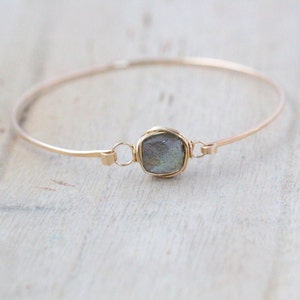 Op de afbeelding: Een delicate goudkleurige armband met een vierkante, iriserende edelsteen in het midden. De edelsteen is gevat in een draadomgeving en de armband heeft een eenvoudig, elegant ontwerp. De armband wordt getoond op een lichtgekleurd oppervlak.