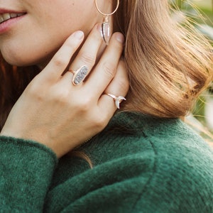 K&ouml;nnte beinhalten: Eine Frau tr&auml;gt einen gr&uuml;nen Pullover mit zwei Ringen an ihren Fingern. Der Ring an ihrem Zeigefinger ist ein goldener Ring mit einem grauen Stein. Der Ring an ihrem kleinen Finger ist ein silberner Ring mit einem wei&szlig;en Stein.