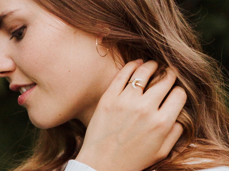 K&ouml;nnte beinhalten: Ein goldener Ring mit einem wei&szlig;en Halbmond-Design. Der Ring wird am Zeigefinger einer Hand getragen.