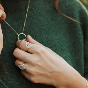 K&ouml;nnte beinhalten: Eine goldene Halskette mit einem runden Anh&auml;nger und einem Halbmond-Anh&auml;nger. Die Kette wird von einer Person mit einem gr&uuml;nen Pullover getragen. Die Person tr&auml;gt auch zwei goldene Ringe an ihrem Finger.