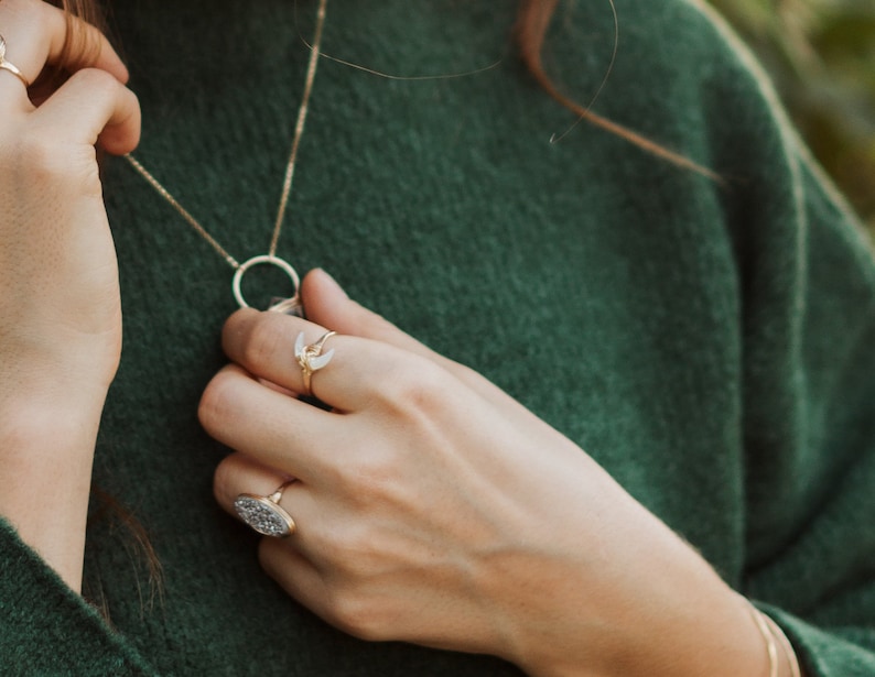 K&ouml;nnte beinhalten: Eine goldene Halskette mit einem runden Anh&auml;nger und einem Halbmond-Anh&auml;nger. Die Kette wird von einer Person mit einem gr&uuml;nen Pullover getragen. Die Person tr&auml;gt auch zwei goldene Ringe an ihrem Finger.