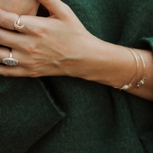 K&ouml;nnte beinhalten: Nahaufnahme einer Hand, die zwei goldene Ringe tr&auml;gt. Der Ring am Zeigefinger ist ein Halbmond-Design. Der Ring am Mittelfinger ist ein gro&szlig;er ovaler Ring mit einer strukturierten Oberfl&auml;che. Die Hand tr&auml;gt ein goldenes Armreif-Armband mit einem kleinen Perlen-Anh&auml;nger.