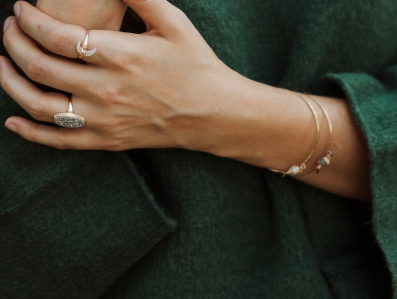 K&ouml;nnte beinhalten: Nahaufnahme einer Hand, die zwei goldene Ringe tr&auml;gt. Der Ring am Zeigefinger ist ein Halbmond-Design. Der Ring am Mittelfinger ist ein gro&szlig;er ovaler Ring mit einer strukturierten Oberfl&auml;che. Die Hand tr&auml;gt ein goldenes Armreif-Armband mit einem kleinen Perlen-Anh&auml;nger.