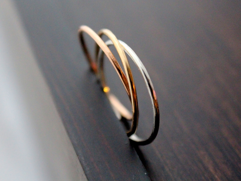May include: Three thin metal rings, one gold, one silver, and one rose gold, are intertwined and stacked on top of each other. The rings are on a dark wooden surface.