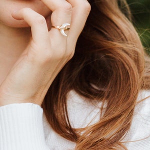 K&ouml;nnte beinhalten: Ein goldener Ring mit einem wei&szlig;en Halbmond-Design. Der Ring wird am Zeigefinger einer Person mit langen braunen Haaren getragen.