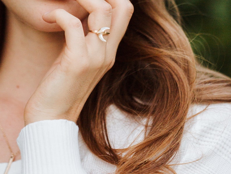 K&ouml;nnte beinhalten: Ein goldener Ring mit einem wei&szlig;en Halbmond-Design. Der Ring wird am Zeigefinger einer Person mit langen braunen Haaren getragen.