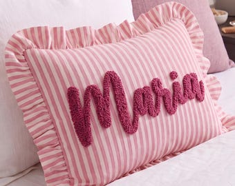 Puede incluir: Un cojín rectangular con un patrón de rayas verticales rosas y blancas y un borde con volantes. El nombre Maria está bordado en el centro con hilo texturizado y copetudo de color rosa oscuro en letra cursiva.