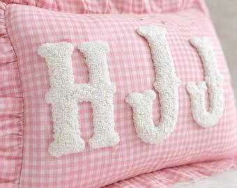 Puede incluir: Cojín rosa de cuadros vichy con letras blancas "HJU". El cojín tiene un borde con volantes y está diseñado para uso decorativo. Las letras en relieve añaden un elemento táctil al diseño.
