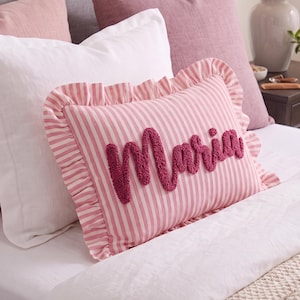 May include: A rectangular throw pillow featuring a pink and white vertical striped pattern with a ruffled border. The name Maria is embroidered in the center using a dark pink, textured, tufted yarn in a cursive script font style.