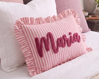 Puede incluir: Un cojín rectangular con un patrón de rayas verticales rosas y blancas y un borde con volantes. El nombre Maria está bordado en el centro con hilo texturizado y copetudo de color rosa oscuro en letra cursiva.