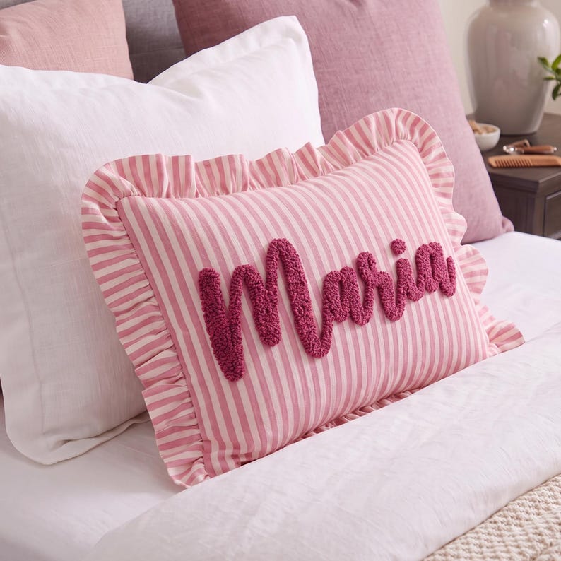 A pink and white striped pillow with the name "Marie" written on it, placed on a bed with other pillows and bedding.