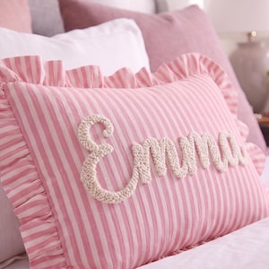 A pink and white striped pillow with the name "Emma" embroidered on it.