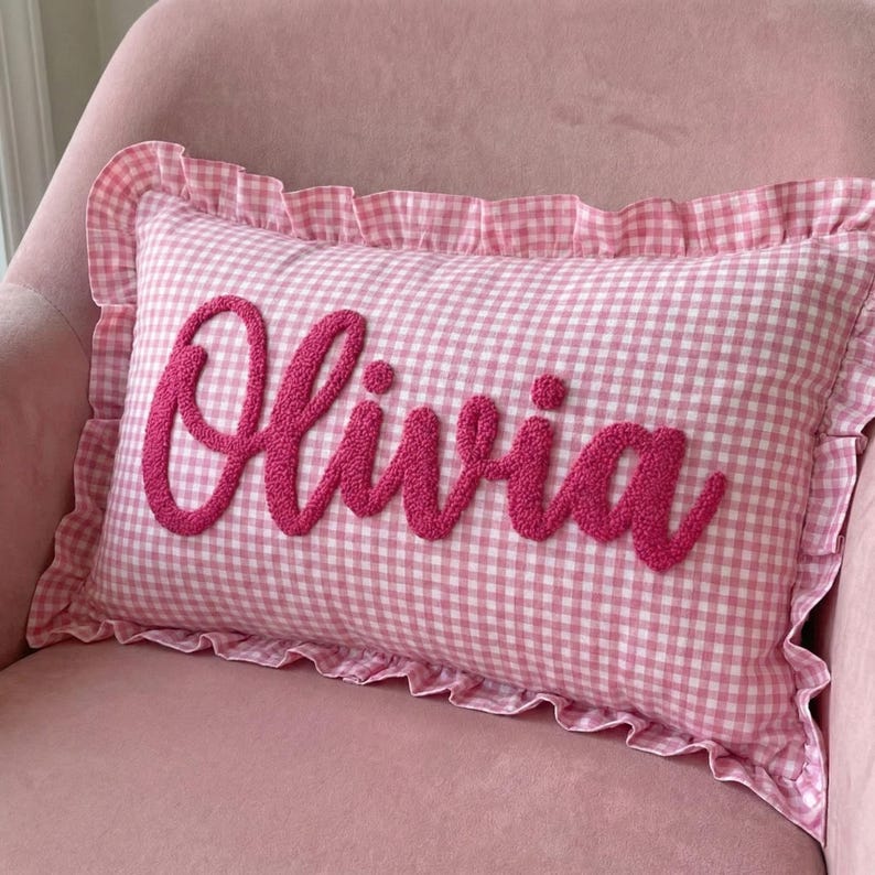 A pink and white gingham pillow with the name "Olivia" embroidered on it.