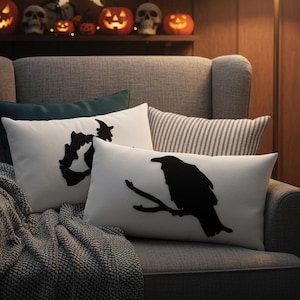 May include: Two white rectangular throw pillows with black Halloween-themed designs rest on a gray armchair. One pillow features a black raven perched on a branch, and the other has a black witch hat design. A gray blanket is draped over the arm.
