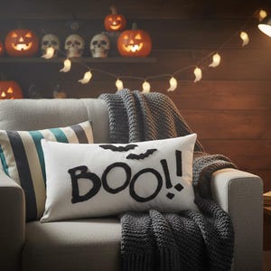 May include: A grey sofa with a white rectangular cushion featuring the word "BOO!!" in black and two bat silhouettes. A grey knitted throw and two decorative cushions are also on the sofa. Lit pumpkins and skulls are on a shelf in the background.