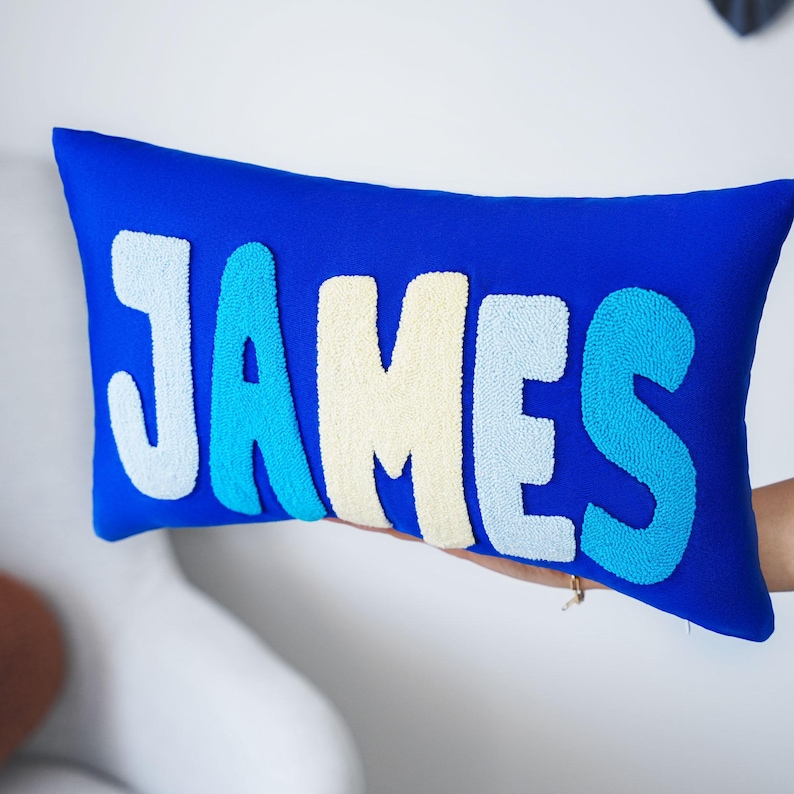 A person is holding a blue pillow with the name "James" embroidered on it.