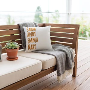 A wooden bench with a white cushion and a gray throw blanket, along with a potted succulent plant, is situated in front of a window overlooking a garden.