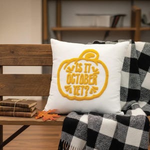 May include: White square pillow with a yellow embroidered pumpkin design and the text "IS IT OCTOBER YET?". The pillow is on a wooden bench with a black and white checkered blanket. A stack of books and a fall leaf are also on the bench.