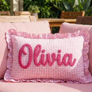 A pink and white gingham pillow with the name "Olivia" embroidered on it, placed on a pink cushion on a wooden chair.