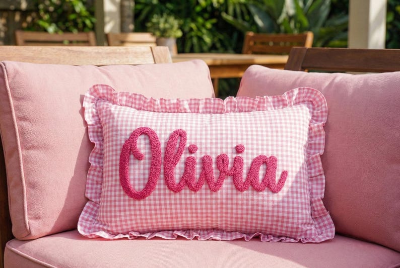 A pink and white gingham pillow with the name "Olivia" embroidered on it, placed on a pink cushion on a wooden chair.