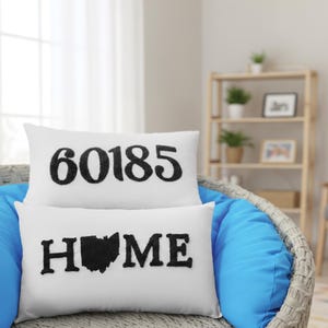 Könnte beinhalten: Zwei weiße, rechteckige Kissen mit schwarzem, gesticktem Text. Das obere Kissen zeigt die Nummer "60185". Das untere Kissen zeigt "HOME" mit einer Umrisslinie von Ohio. Die Kissen liegen auf einem blauen Stuhl.