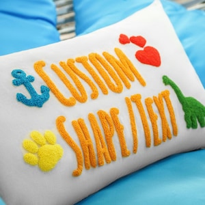 A white pillow with colorful embroidered text and images, including a blue anchor, a red heart, a green dinosaur, and a yellow paw print.