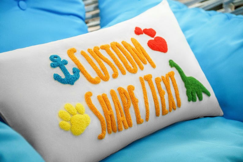 A white pillow with colorful embroidered text and images, including a blue anchor, a red heart, a green dinosaur, and a yellow paw print.