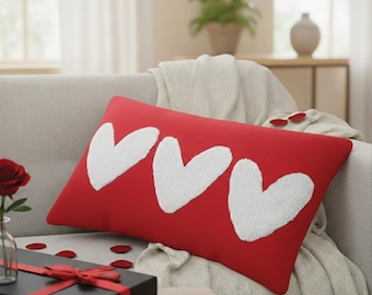 May include: A red rectangular pillow with three white heart shapes. The pillow is on a light gray sofa, with a gift box and rose petals nearby. The pillow is a decorative item.