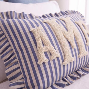 A decorative pillow with the letters "AM" embroidered on it, featuring a striped pattern in shades of blue and white.