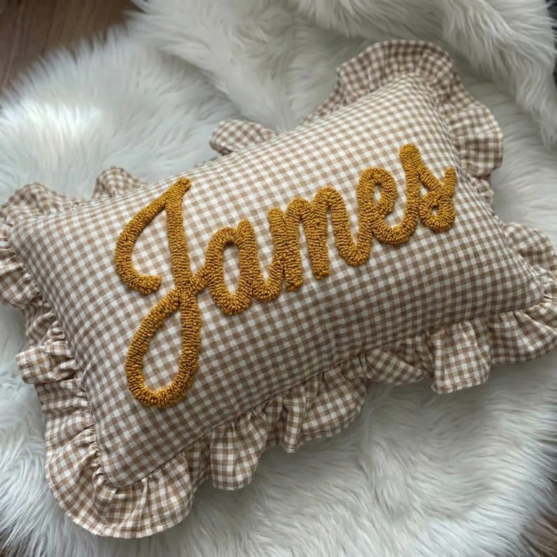 A decorative pillow with the name "James" embroidered on it, placed on a white furry surface.