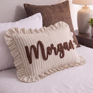 a bed with a decorative pillow featuring the name "Morgan" embroidered on it. The pillow is placed on a white bedspread, and there are other pillows and decorative items in the background.