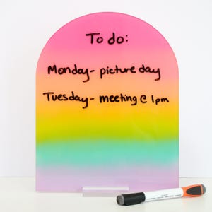 May include: A rainbow-colored acrylic to-do list board with a rounded top. The board has the words "To do:" and the schedule for Monday and Tuesday written in black marker. A white and orange marker with a black tip rests on the surface.