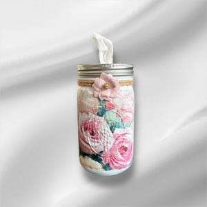 Op de afbeelding: Een decoratieve tissuehouder van een weckpot. De pot is versierd met een bloemenpatroon in roze en wit. Een roze bloem en touw accentueren de pot. Een tissue wordt uit de bovenkant getrokken.