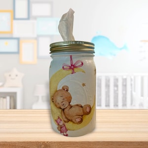 Pode incluir: Um frasco de vidro branco com tampa dourada, decorado com um urso dormindo em uma lua crescente, estrelas e uma fita rosa. O frasco está cheio de lenços de papel e é um porta-lenços bonito e caprichoso.