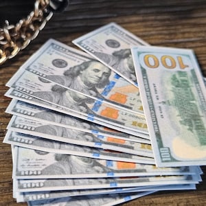 Puede incluir: Una pila de diez billetes de 100 euros con una cadena de oro y un bolso negro en el fondo.