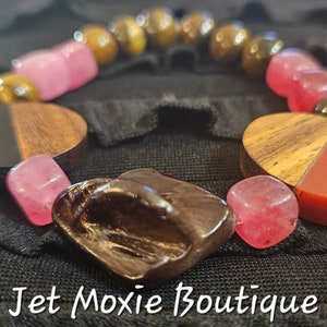 May include: A bracelet made with pink, red, brown, and black beads. The bracelet has a wooden charm with a carved design. The bracelet is on a black fabric background. Jet Moxie Boutique.