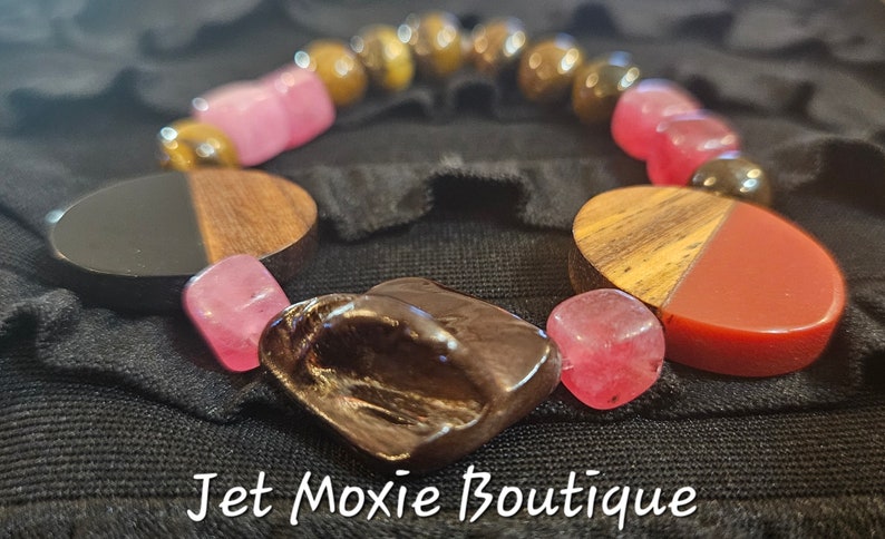May include: A bracelet made with pink, red, brown, and black beads. The bracelet has a wooden charm with a carved design. The bracelet is on a black fabric background. Jet Moxie Boutique.