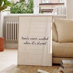 Puede incluir: Una impresión enmarcada con el texto "love's never wasted when it's shared" se apoya en un sofá beige en una habitación luminosa. También se ven una gran planta en maceta y una silla de madera. La habitación tiene una paleta de colores neutros.